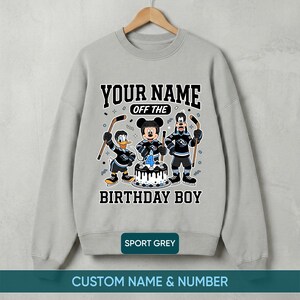 May include: Grey sweatshirt with a hockey-themed birthday design. It features cartoon characters, a birthday cake with a number 4 candle, and the text "YOUR NAME OFF THE BIRTHDAY BOY".