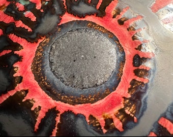Puede incluir: Primer plano abstracto de un diseño circular con un círculo gris central rodeado de anillos concéntricos. El anillo exterior presenta un patrón rojo vibrante con acentos negros y marrones, creando un contraste visual llamativo.