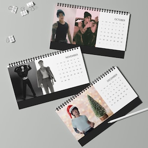 May include: Three spiral-bound desk calendars featuring black and white photographs of a person in various outfits. The pages display the months of October, November, and December with corresponding grids. A white pen rests near the December calendar.