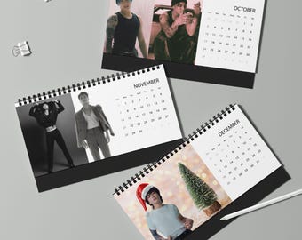 May include: Three spiral-bound desk calendars featuring black and white photographs of a person in various outfits. The pages display the months of October, November, and December with corresponding grids. A white pen rests near the December calendar.