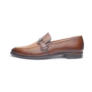 May include: A brown leather loafer featuring a perforated texture across the upper surface. The shoe includes a dark metallic horsebit hardware detail across the vamp, a low heel, and a rounded toe profile, presented from a side view perspective.