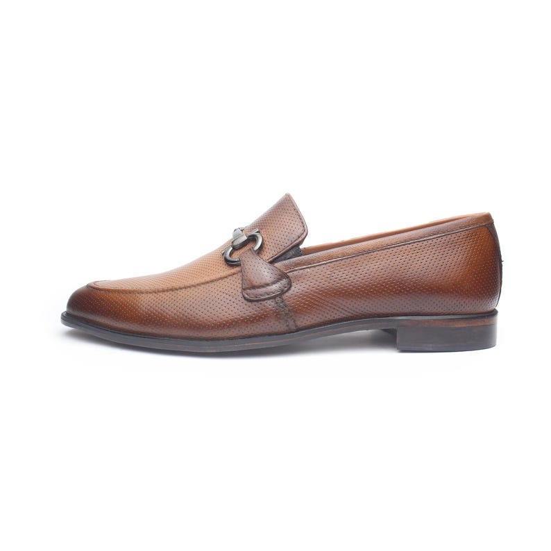 May include: A brown leather loafer featuring a perforated texture across the upper surface. The shoe includes a dark metallic horsebit hardware detail across the vamp, a low heel, and a rounded toe profile, presented from a side view perspective.