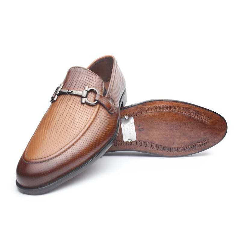May include: A brown leather loafer with a perforated texture and a dark metal bit ornament across the vamp. The shoe is positioned to show the smooth, dark brown sole, which features a small metal plate clearly displaying the size 40.