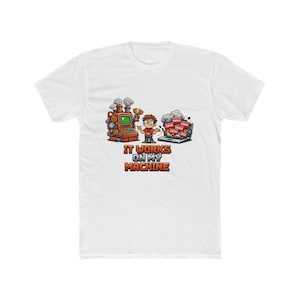 Può includere: T-shirt bianca con grafica colorata. La grafica presenta un cartone animato di una persona che tiene un trofeo, un computer e una macchina a vapore. Il testo "IT WORKS ON MY MACHINE" è in basso.