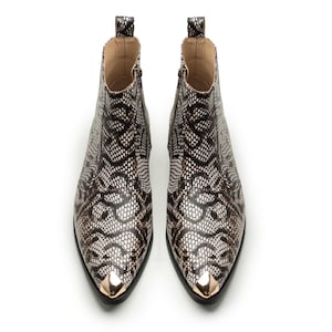 May include: A pair of ankle boots with a snakeskin pattern in black, white, and brown. The boots have pointed toes with gold-colored metal tips and zippers on the sides. The boots are set against a white background.