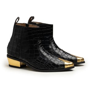 Pode incluir: Um par de botins pretos com um padrão em relevo de crocodilo. Os botins apresentam uma biqueira metálica em tom dourado e um salto de bloco a condizer no mesmo tom dourado metálico, com uma presilha na parte de trás do cano.