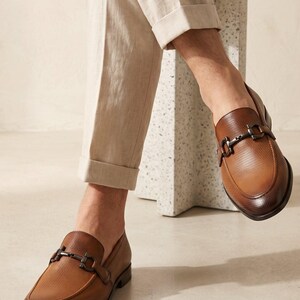May include: A pair of brown leather loafers featuring a perforated texture, a dark brown gradient finish at the toe and heel, and a metallic horsebit detail across the vamp. The shoes are worn with light beige cuffed trousers.