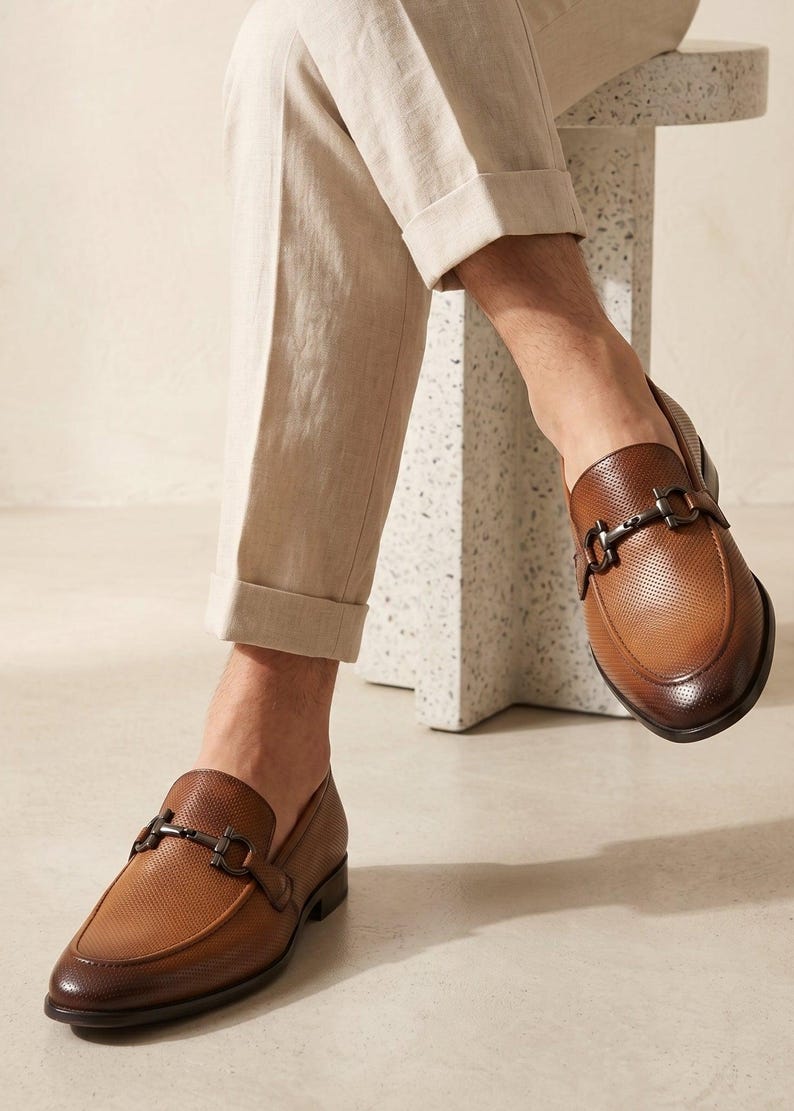 May include: A pair of brown leather loafers featuring a perforated texture, a dark brown gradient finish at the toe and heel, and a metallic horsebit detail across the vamp. The shoes are worn with light beige cuffed trousers.