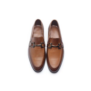 May include: A pair of tan brown loafers featuring a textured, micro-perforated leather upper and a dark metallic horsebit hardware detail across the vamp. These slip-on shoes have a rounded toe, a smooth interior lining, and a low-profile silhouette.