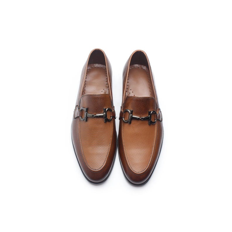 May include: A pair of tan brown loafers featuring a textured, micro-perforated leather upper and a dark metallic horsebit hardware detail across the vamp. These slip-on shoes have a rounded toe, a smooth interior lining, and a low-profile silhouette.