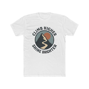 May include: White t-shirt with a circular graphic featuring a mountain, a winding path, and a setting sun. The text "CLIMB HIGHER SHINE BRIGHTER" is printed around the graphic in a bold, sans-serif font. The shirt is made of a soft, comfortable material.