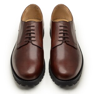 Peut inclure: Paire de chaussures en cuir marron avec des lacets marron foncé. Les chaussures ont un bout arrondi et une semelle épaisse et noire. Présentées sur fond blanc.