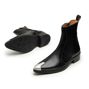 May include: A pair of black leather ankle boots featuring a pointed toe with a metallic silver cap, a side zipper closure, a pull tab at the heel, and a black sole with a circular tread pattern visible on the bottom of the second boot.