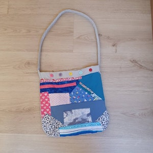 Peut inclure: Un sac à bandoulière patchwork avec une sangle grise. Le sac présente une variété de motifs de tissus, notamment des cœurs, des imprimés floraux et des couleurs unies. Le bord supérieur est orné de boutons colorés. Le sac a une forme rectangulaire avec un motif en forme de fenêtre.