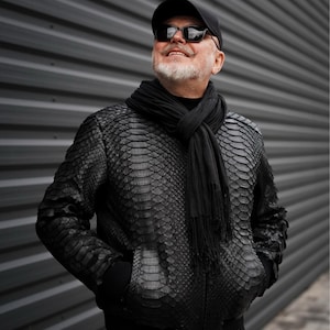 Może przedstawiać: Mężczyzna w czarnej skórzanej kurtce bomber z czarnym szalikiem i okularami przeciwsłonecznymi. Stoi przed szarą falistą ścianą metalową.