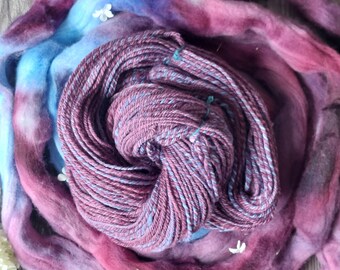May include: A skein of hand-dyed yarn in shades of purple, blue, and pink. The yarn is wrapped around a bundle of hand-dyed fiber in similar colors.