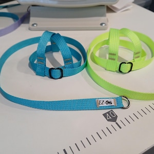 May include: A bright blue and a neon yellow nylon pet harness and leash set. The blue harness features a small white fabric tag with the black text EZ-On. Both harnesses have black plastic adjustment buckles and a metal D-ring for leash attachment.