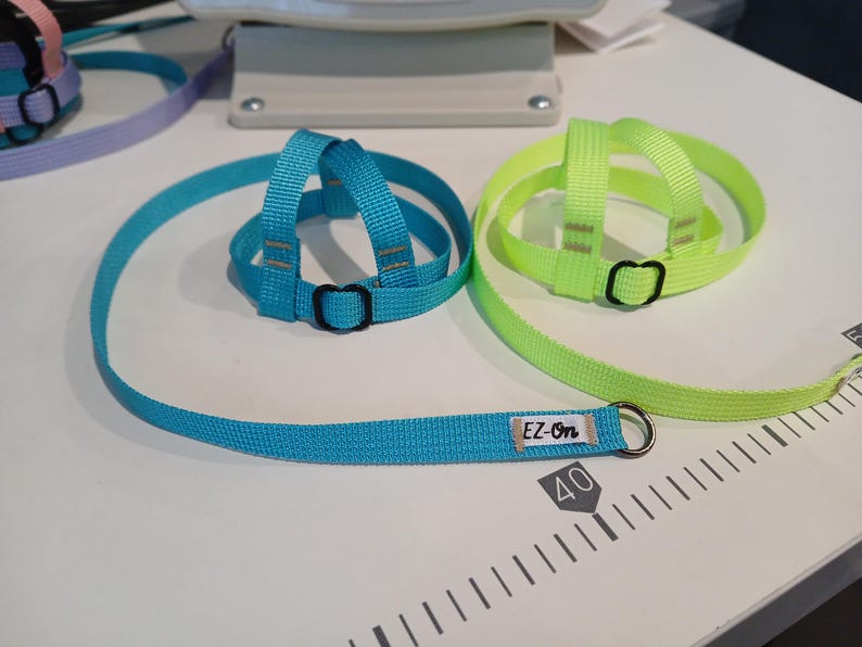 May include: A bright blue and a neon yellow nylon pet harness and leash set. The blue harness features a small white fabric tag with the black text EZ-On. Both harnesses have black plastic adjustment buckles and a metal D-ring for leash attachment.
