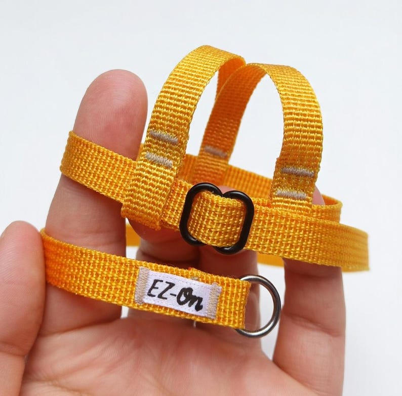 May include: A bright yellow nylon pet harness featuring a woven texture, a black plastic adjustable slider, and a silver metal D-ring. A small white fabric tag is sewn onto the strap with the black text EZ-On printed in a cursive font.
