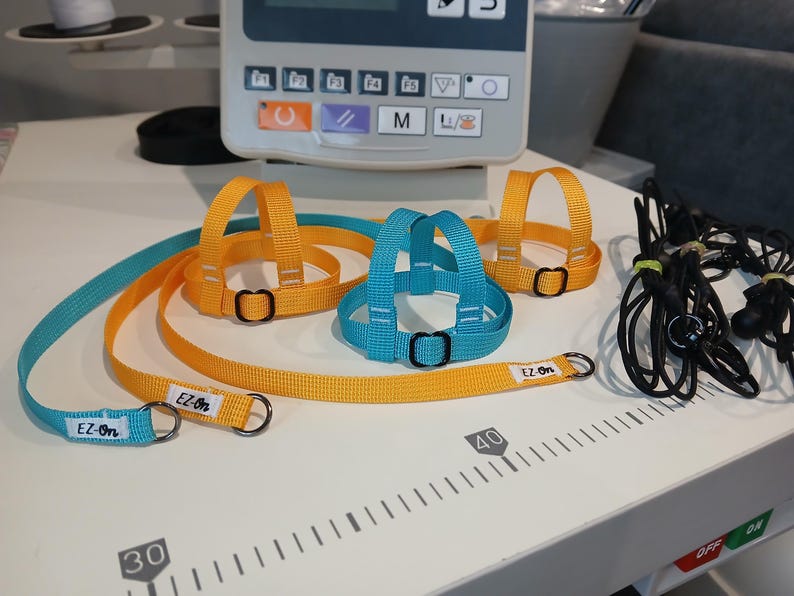 May include: Two nylon pet harnesses with matching leashes in bright yellow and turquoise. Each harness features adjustable black metal sliders and a white tag labelled EZ-On. The leashes include metal D-rings and are positioned on a scale.