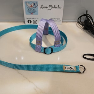 May include: A turquoise and lavender woven nylon bird harness featuring a black plastic buckle and a long turquoise leash with a metal ring. A small white tag with the text EZ-On is attached to the leash. A separate black cord with a metal clip.