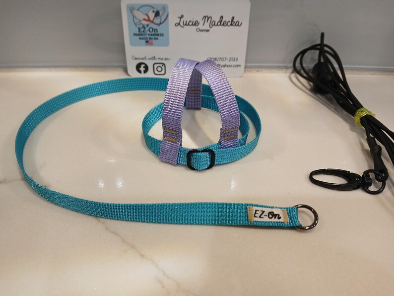 May include: A turquoise and lavender woven nylon bird harness featuring a black plastic buckle and a long turquoise leash with a metal ring. A small white tag with the text EZ-On is attached to the leash. A separate black cord with a metal clip.