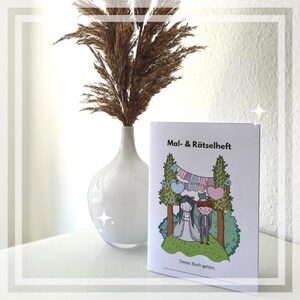May include: A white booklet with the title "Mal- & R&auml;tselheft" and a colorful illustration of a bride and groom standing under a floral archway. The text "Dieses Buch geh&ouml;rt." is at the bottom of the page.