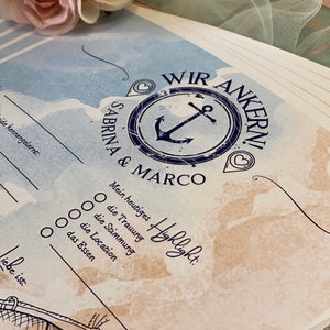 May include: A nautical-themed wedding guest book with the words "WIR ANKERN!" and the names "Sabrina & Marco" in a navy blue anchor design. The book has fill-in-the-blank sections and a watercolor background with a soft pink and cream flower.