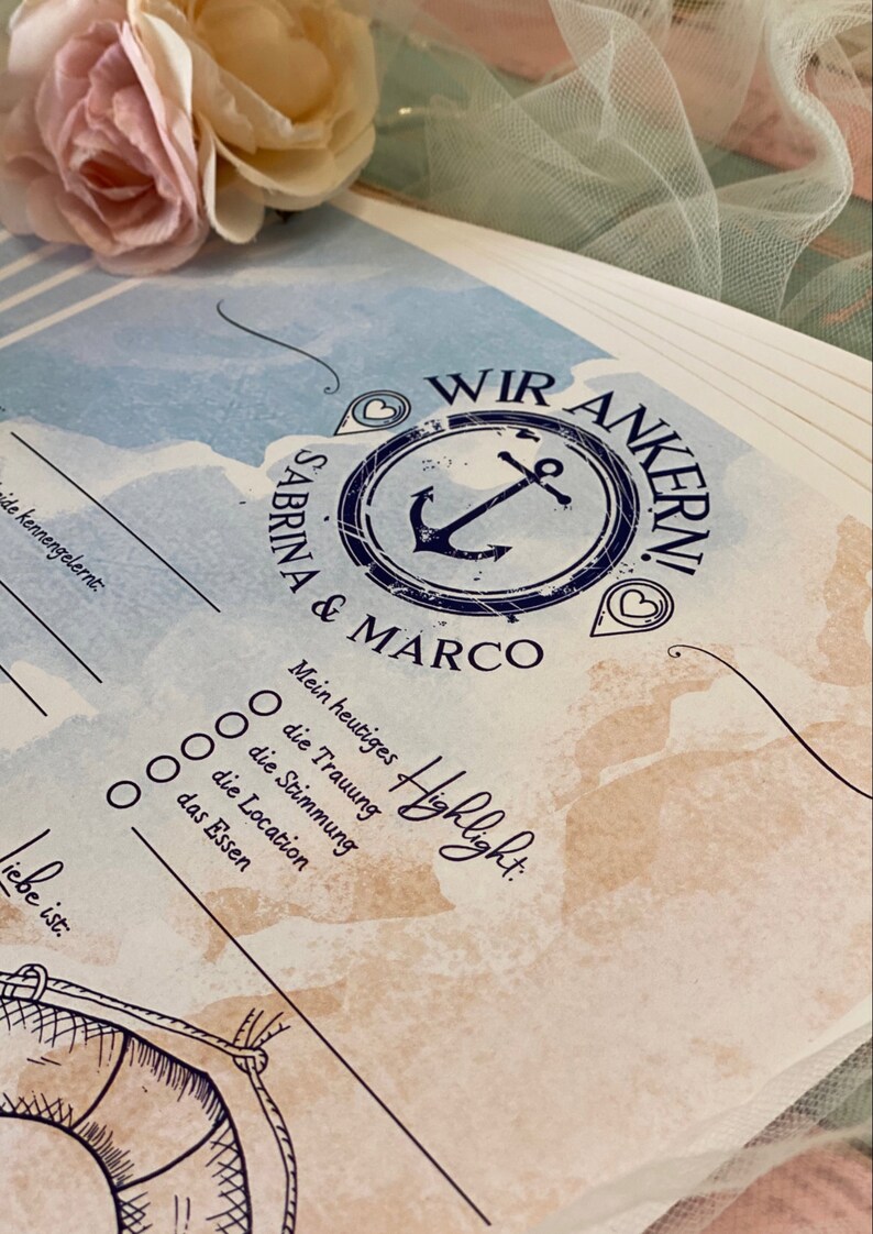 May include: A nautical-themed wedding guest book with the words "WIR ANKERN!" and the names "Sabrina & Marco" in a navy blue anchor design. The book has fill-in-the-blank sections and a watercolor background with a soft pink and cream flower.