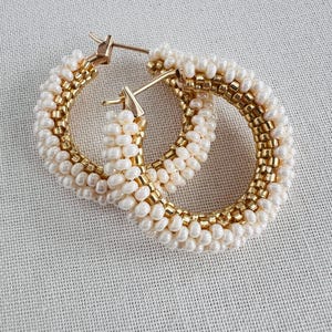 May include: A pair of hoop earrings featuring a design of white pearls and gold beads. The earrings have a gold-colored post closure. The pearls and beads are arranged in concentric circles, creating a textured appearance.