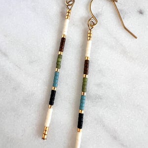 May include: Dangling earrings with a linear design. The earrings feature a gold-colored hook and are composed of small, colorful beads in white, brown, green, blue, and black. The beads are separated by gold-colored spacers.