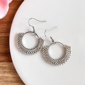 May include: A pair of silver hoop earrings with a beaded design. The earrings are made of small, shiny beads arranged in a semi-circular pattern around a silver hoop. The earrings are displayed on a white surface.