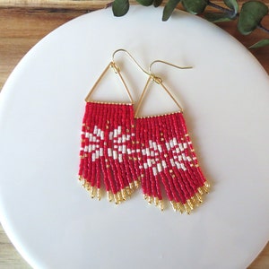 May include: A pair of red and white beaded earrings with a gold triangle frame. The earrings feature a snowflake pattern and dangle from gold hooks.