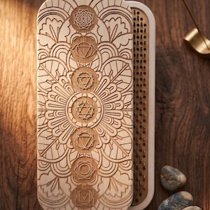 May include: A light-colored wooden box with a carved mandala design and chakra symbols. The box is open, revealing a pegboard interior. Incense burns nearby, and small stones are scattered around.