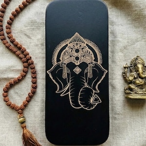 May include: A black rectangular box featuring a gold illustration of an elephant-headed deity. A brown beaded necklace and a small bronze statue of the same deity are also present. The items are on a neutral-coloured fabric.