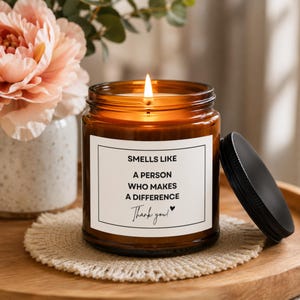 May include: A lit candle in an amber glass jar with a black lid. The white label features black text that reads: SMELLS LIKE A PERSON WHO MAKES A DIFFERENCE Thank you! with a small heart icon. The candle sits on a round, fringed, woven coaster.