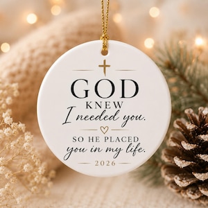 May include: A round white ceramic ornament featuring a gold cross and the black text: GOD KNEW I needed you. SO HE PLACED you in my life. 2026. The ornament includes a small gold heart icon and is suspended by a thin gold cord for hanging purposes.