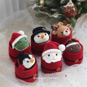 Puede incluir: Seis figuras navideñas de ganchillo en recipientes rojos, incluyendo una criatura verde, un muñeco de nieve, un reno, un pingüino, Santa Claus y un personaje marrón. Cada recipiente tiene un cinturón negro con hebilla dorada. Texto: @lulupetitedoll.