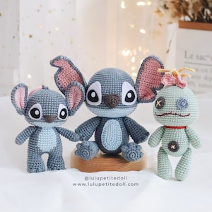 Może przedstawiać: Trzy pluszowe zabawki wykonane na szydełku: niebieski Stitch z różowymi uszami, mniejszy niebieski Stitch z różowymi uszami i zielony Eksperyment 626 z różową kokardką. Zabawki siedzą na drewnianej platformie. Obraz zawiera tekst "@luluptitedoll www.luluptitedoll.com".