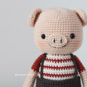 May include: A crocheted pig wearing a red and white striped shirt with brown overalls. The pig has black eyes and a pink snout.