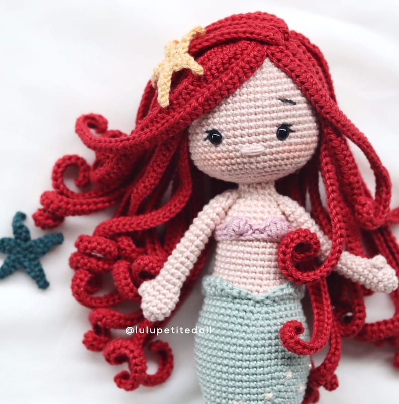 May include: A crocheted mermaid doll with long red hair, a pink top, and a green tail. The doll is wearing a starfish in her hair.