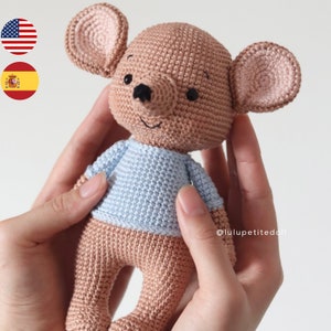 Puede incluir: Un juguete de ratón de crochet con pelaje marrón, una camisa azul claro y ojos negros. El ratón está siendo sostenido por dos manos.