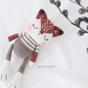 Puede incluir: Un juguete de zorro de crochet con rayas marrones, blancas y grises. El zorro tiene los ojos cerrados y una sonrisa cosida. El juguete está acostado sobre una superficie blanca. @luluptitedoll