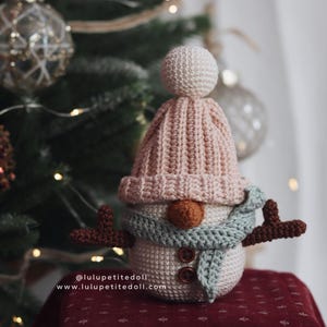 May include: Crocheted snowman figurine with a light pink hat, a light blue scarf, and brown button eyes and nose. The snowman has brown antlers and a white body. The website address is visible on the bottom left.