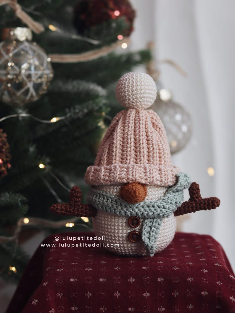 May include: Crocheted snowman figurine with a light pink hat, a light blue scarf, and brown button eyes and nose. The snowman has brown antlers and a white body. The website address is visible on the bottom left.