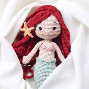 May include: A crocheted mermaid doll with long red hair, a light pink seashell bra, and a teal tail. The doll is lying on a white fabric background.