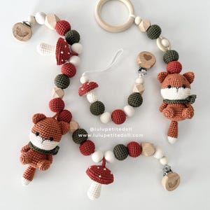 May include: A collection of crocheted baby accessories with a fox and mushroom design. The set includes a pacifier clip, a wooden ring, and beaded garlands. The colour scheme features brown, red, green, and white. The website address is visible in the image.