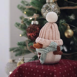 May include: A crocheted snowman ornament with a light pink hat and a light blue scarf. The snowman has brown twig arms and button eyes. The background includes a Christmas tree with ornaments. The text "@lulupetitedoll www.lulupetitedoll.com" is visible.