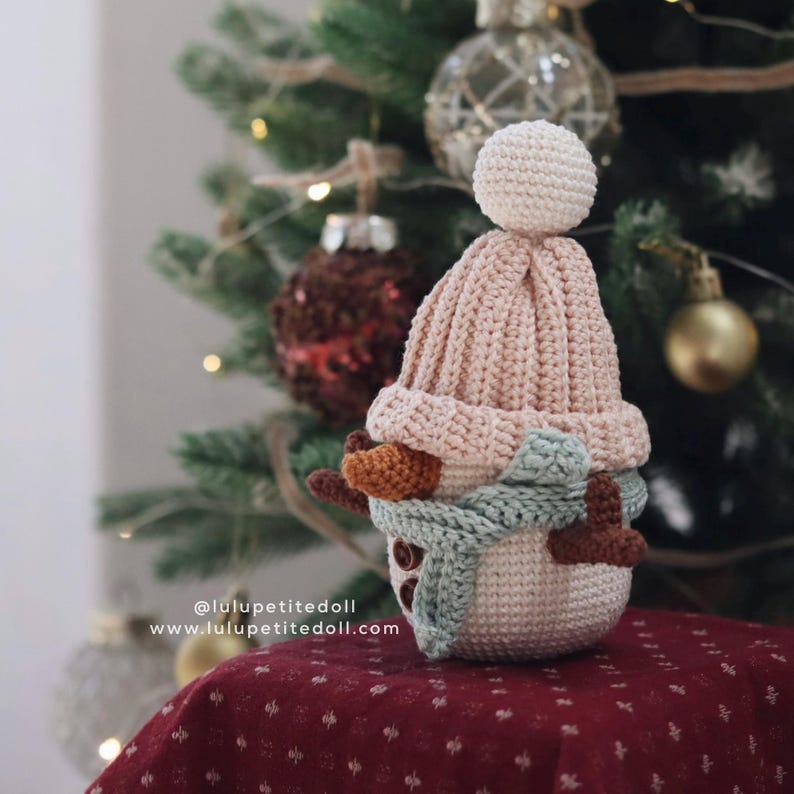 May include: A crocheted snowman ornament with a light pink hat and a light blue scarf. The snowman has brown twig arms and button eyes. The background includes a Christmas tree with ornaments. The text "@lulupetitedoll www.lulupetitedoll.com" is visible.