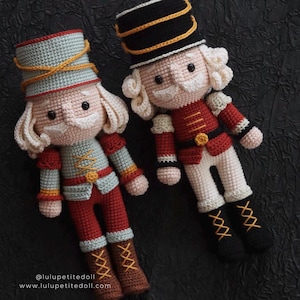 May include: Two crocheted nutcracker dolls, one with a blue and gold hat and the other with a black and gold hat. Both dolls are wearing red and white uniforms with gold accents. The dolls are standing on a black background.